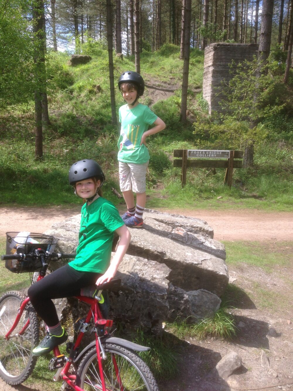 A girl and boy resting next to a big rock in the forest, wearing bike helmets and smiling at the camera