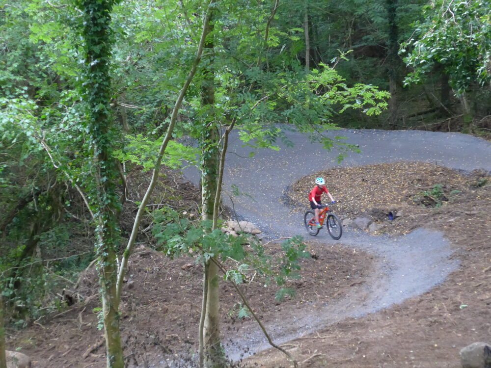 A boy seen from the front, at a distance, weaving his way around some big berms on a mountain bike