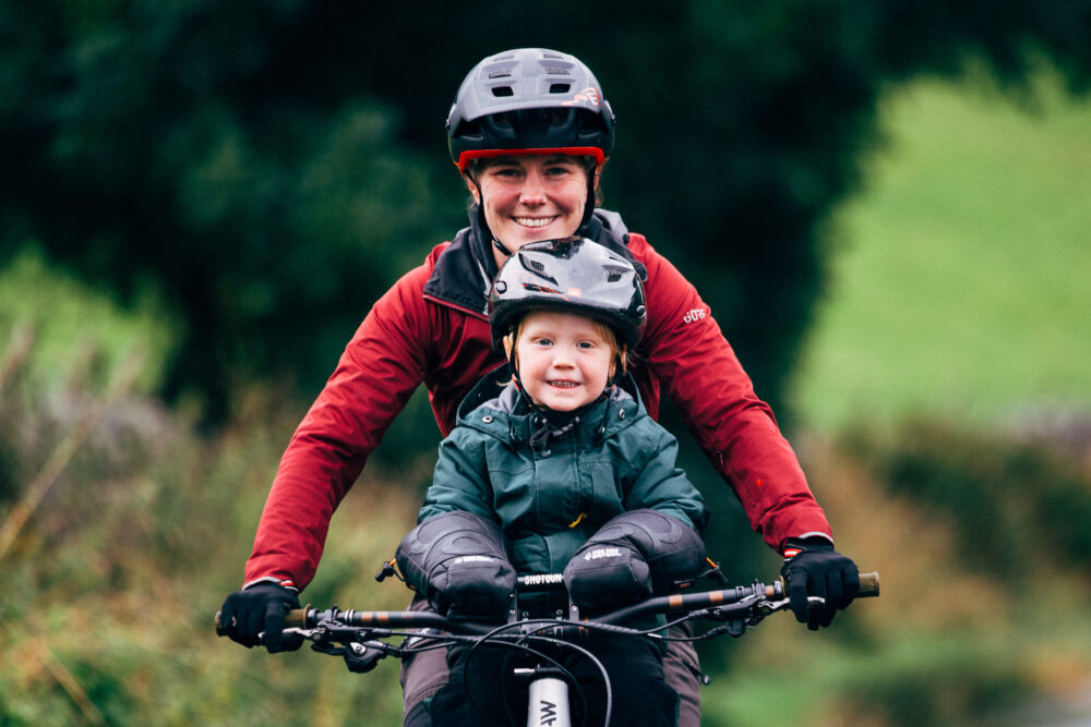 Kids Ride Shotgun Pogies for winter riding wtih toddlers Small toddler on front bike seat keeping his little hands warm in the pogies. He has a big smile on his face, and is mum is behind him steering the bike,