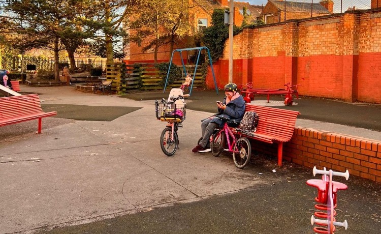 Taking a breather Cycling in Ramadan - A Muslim woman wearing a Hijab under her bike helmet, checking her phone on a park bench surrounded by her family's bikes
