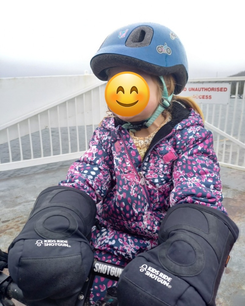 Girl in a blue helmet and flowery jacket using the Kids ride shotgun pogies while on a bike on board a boat.