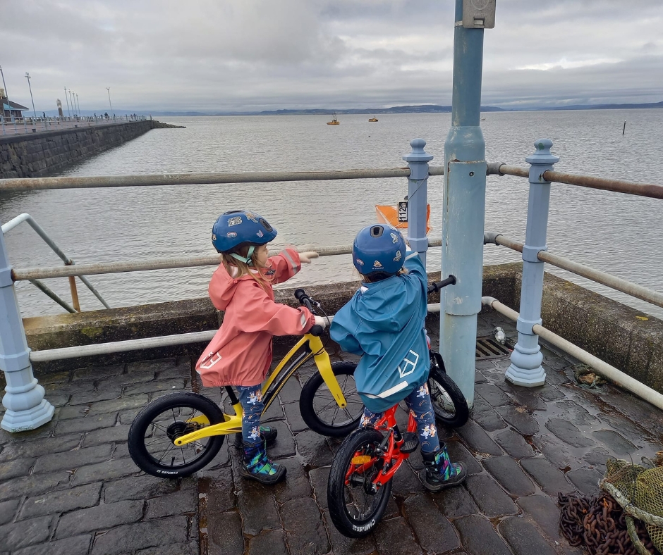 two girls on balance bikes pointing at a sign next to the sea