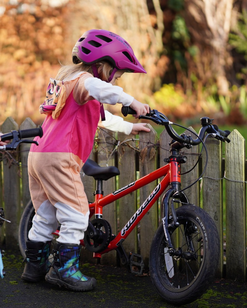 Bikes for girls: A little girl wearing all pink and a pink mountain bike helmet is propping her red bike against a wall