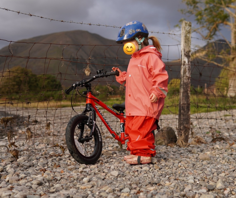 A girl in a pink coat stadning on a pebbly beach next to the kidvelo rookie 14 bike to review