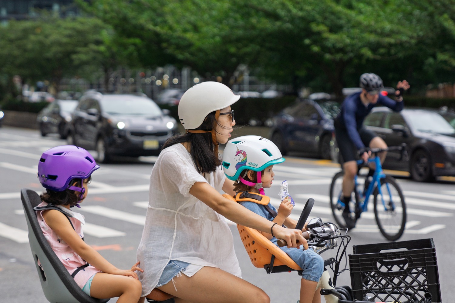 An Asian woman cycling a bike with her toddler on a front-mounted bike seat, wearing an Ozmo e-bike helmet for babies, and a small child on a rear-mounted bike seat, also wearing an Ozmo helmet.