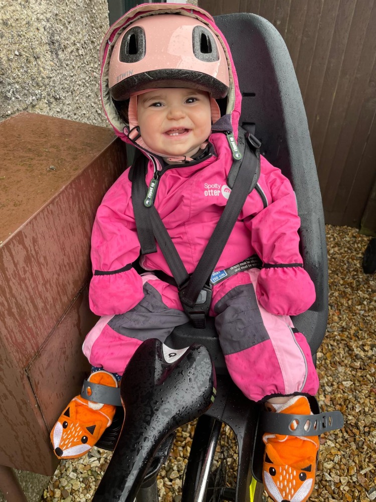 girl in a pink onesie and pink helmet smiling on a rear bike sear