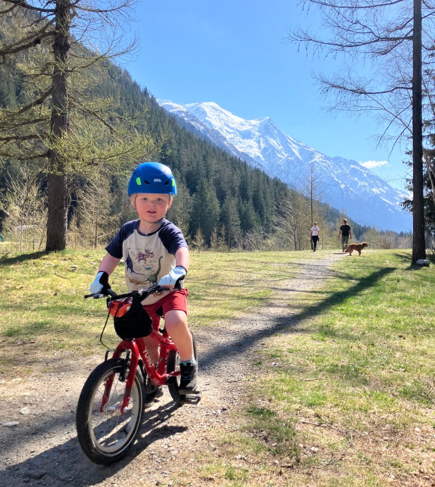 A little boy riding his red yomo bike with a blue helmet on a gravel path on a sunny day with mountains behind