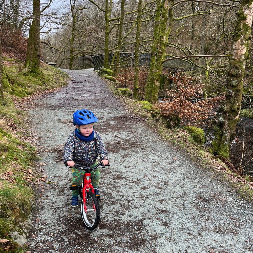 A little boy riding his red yomo bike with a blue helmet and blue waterproof onsie on a drizzy day