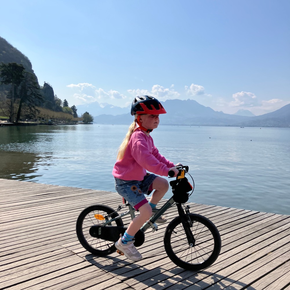 BTWIN 16 Discover 900 review- a girl in a pink jumper riding next to a very blue lake with big mountains in the background