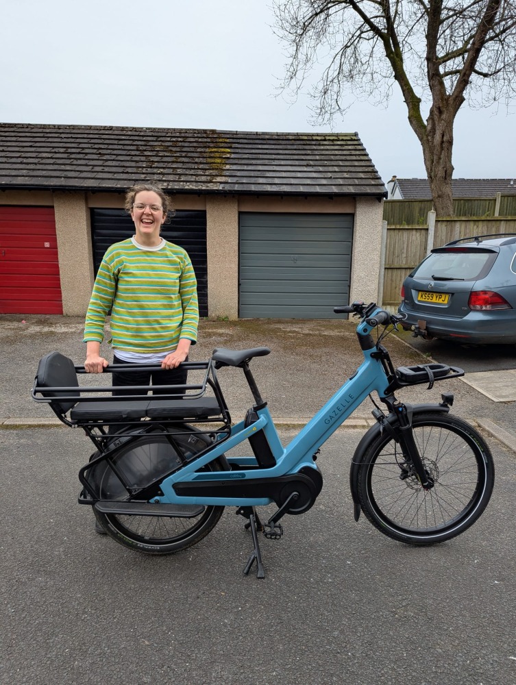 Emily, in a green stripy top standing with the blue Gazelle Cabby longtail bike
