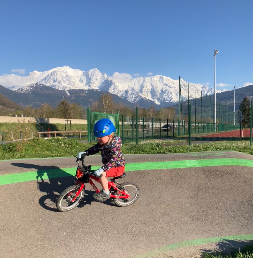 A little boy riding his red yomo bike with a blue helmet on a pump track with big snowy mountains behind him on a sunny day