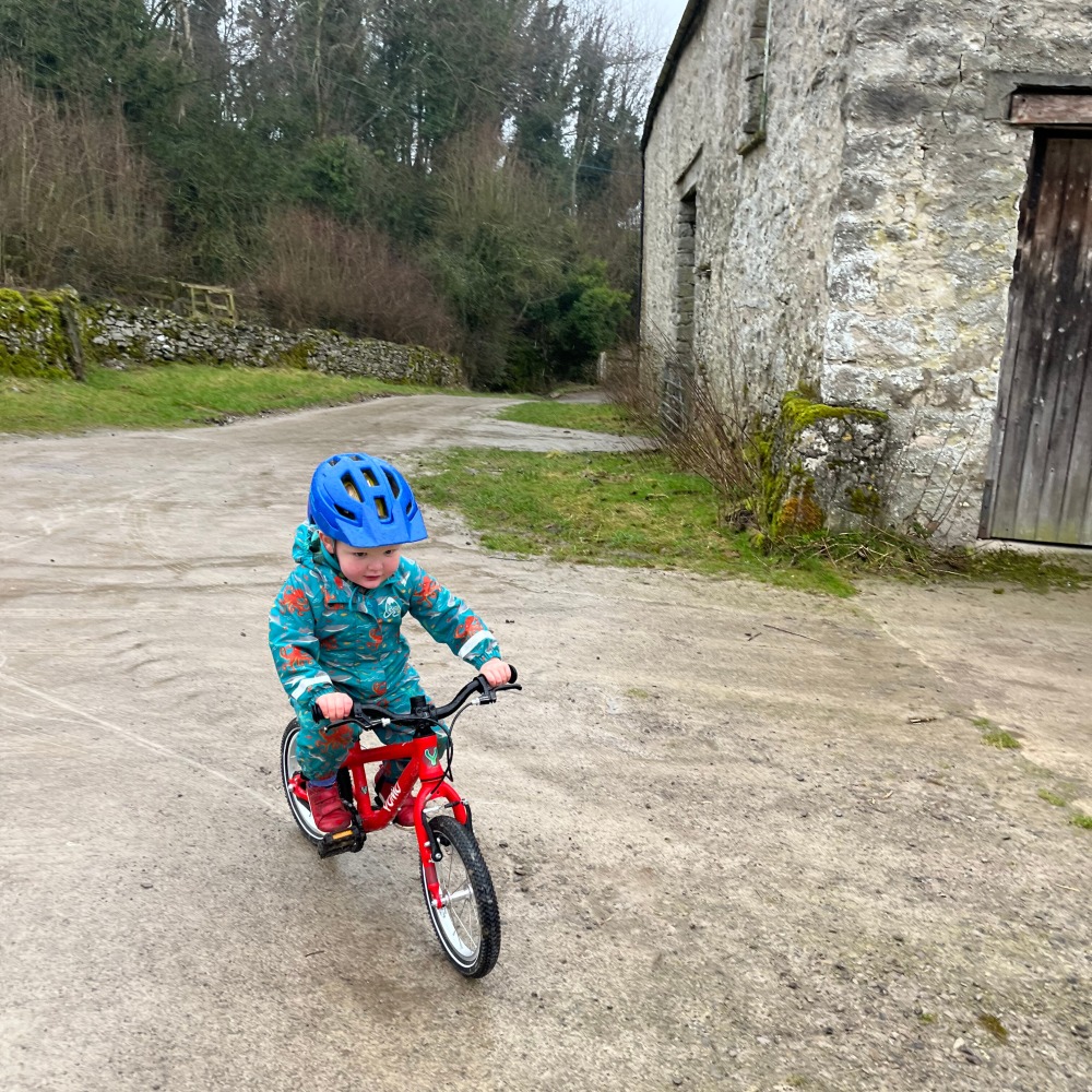 A little boy riding his red yomo bike with a blue helmet and blue waterproof onsie on a drizzy day