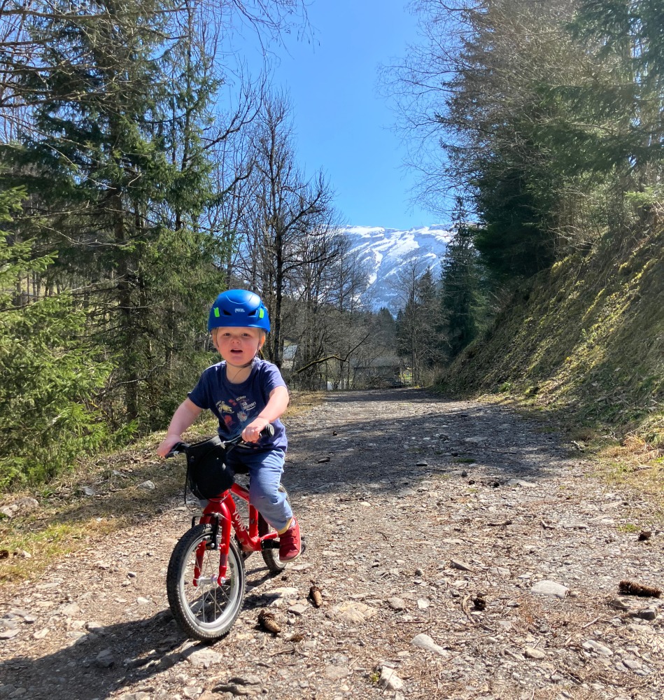 A little boy riding his red yomo bike with a blue helmet on a gravel path on a sunny day with mountains behind