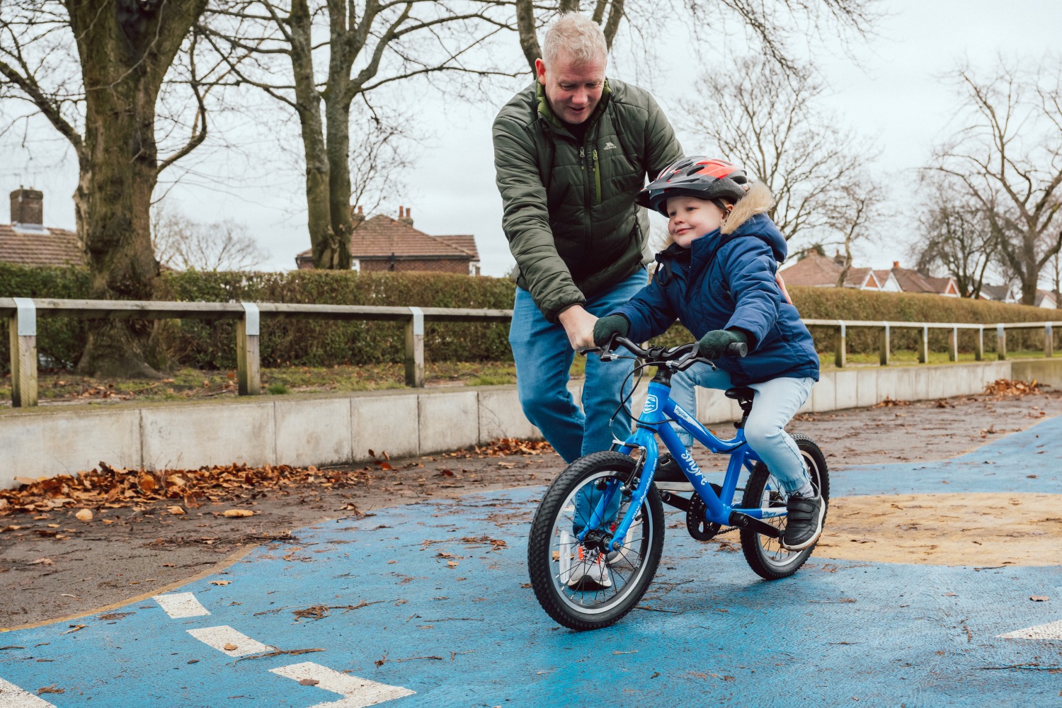 A little boy pedalling a blue Shyre bikes Hopton 16 in the park with his dad holding onto him and running alongside