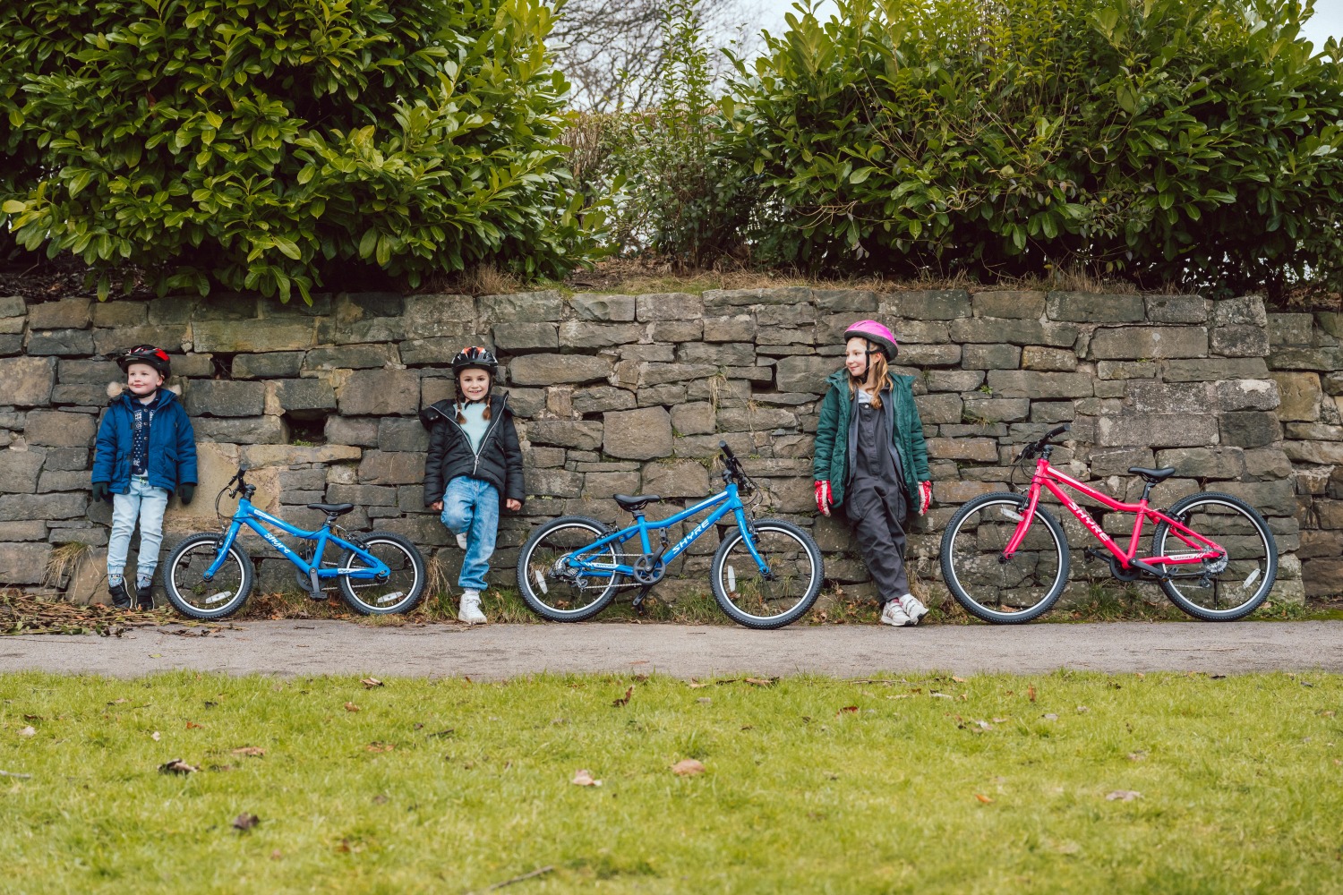 Three kids leaning against a brick wall, with their shyre bikes next to them
