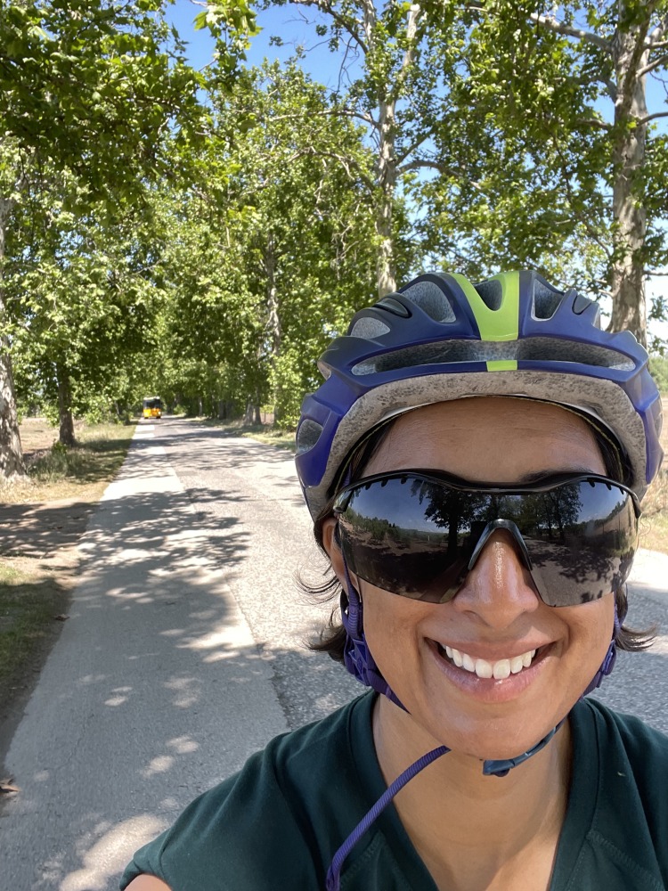 A selfie of Sahir smiling while cycle touring, in her helmet and sunglasses, with a white gravel path and trees behind her
