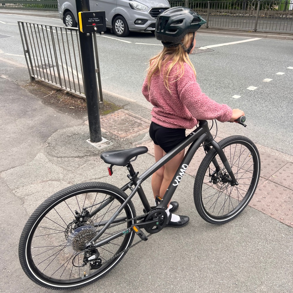 YOMO 24 review- a girl in a pink jumper and yellow helmet is waiting at some traffic lights