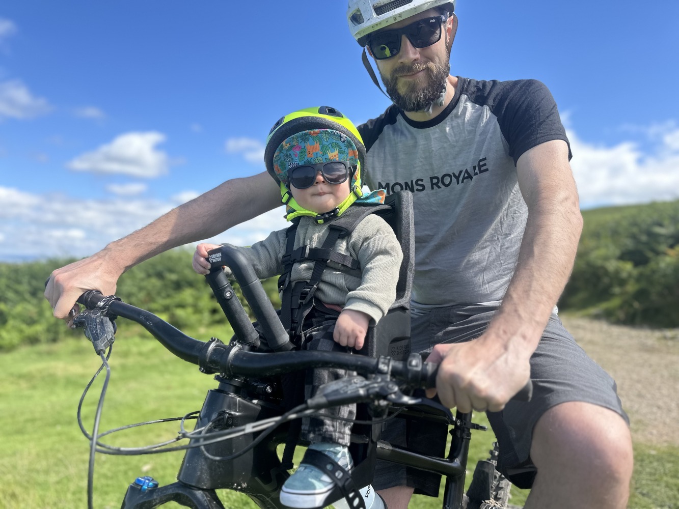A little boy with a yellow helmet and sunglasses smiling on the kids ride shotgun pro evo seat with his dad