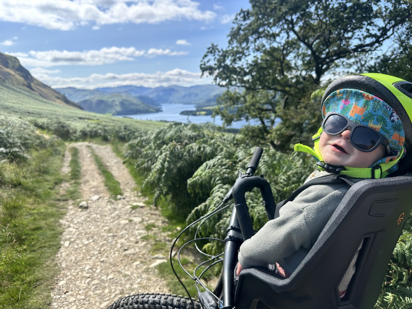 A little boy with a yellow helmet and sunglasses smiling on the kids ride shotgun pro evo seat with a lake and mountains begind him