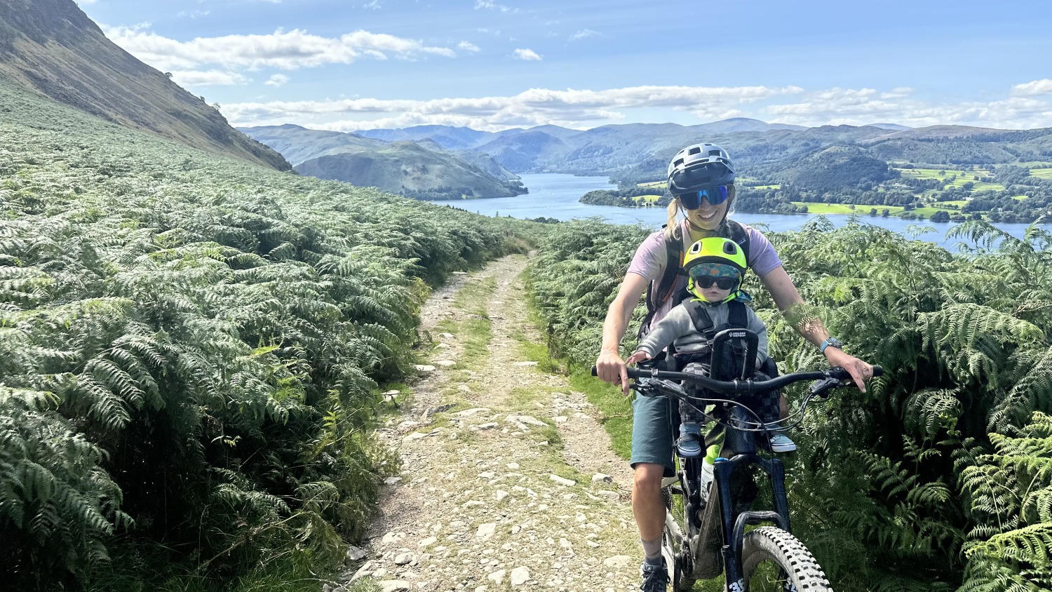 A little boy with a yellow helmet and sunglasses smiling on the kids ride shotgun pro evo seat with his mum, with lake ullswater and mountains behind them