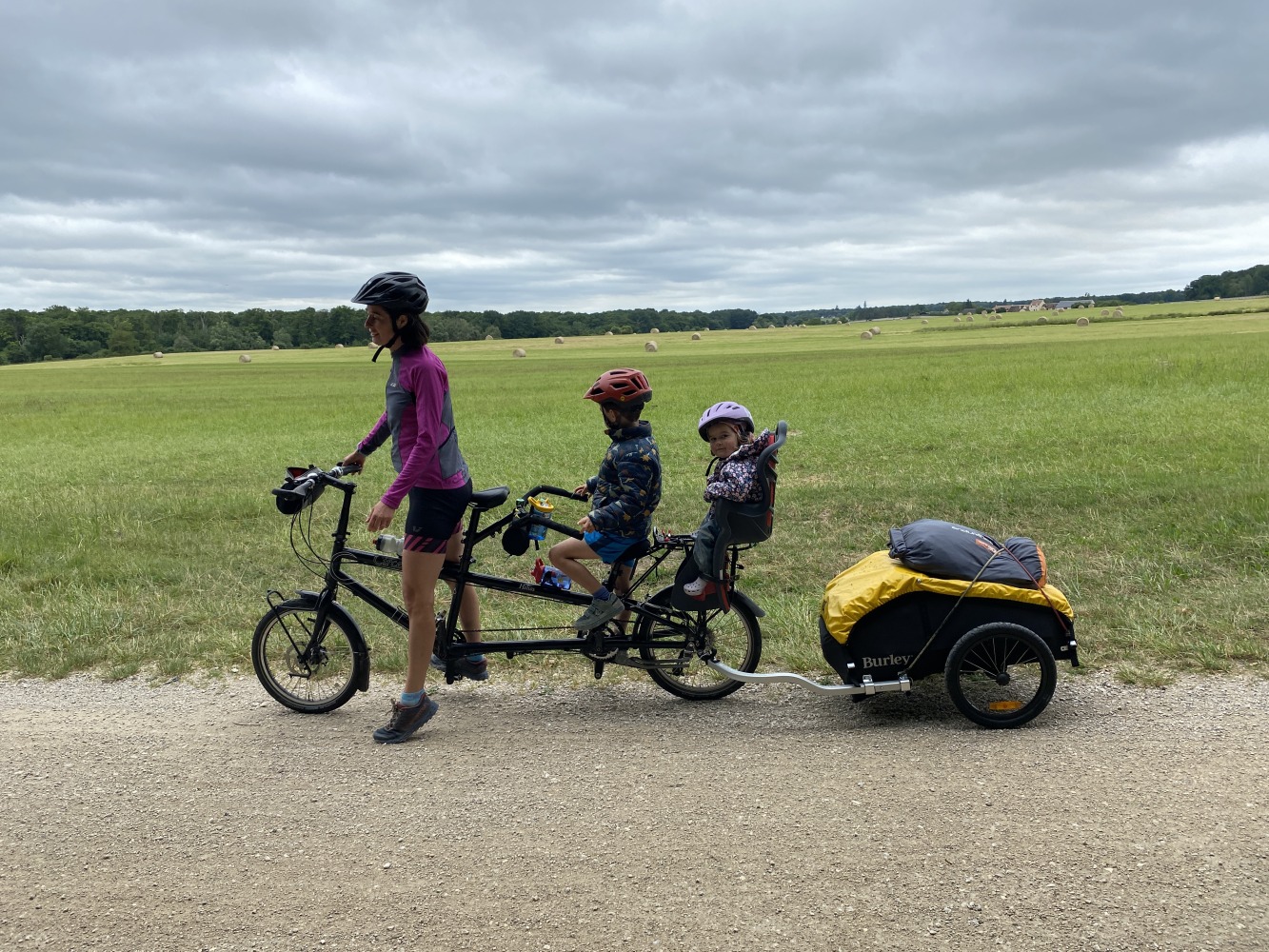 the yellow and black Burley Nomad cargo trailer hitched to a tandem with a mum, son and baby on board smiling infront of a green field