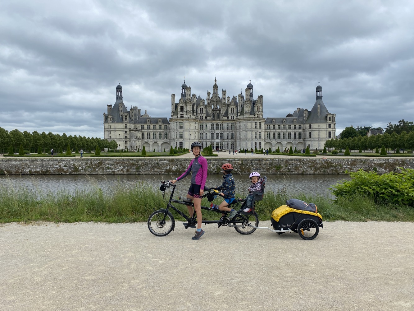 the yellow and black Burley Nomad cargo trailer hitched to a tandem with a mum, son and baby on board smiling infront of a castle