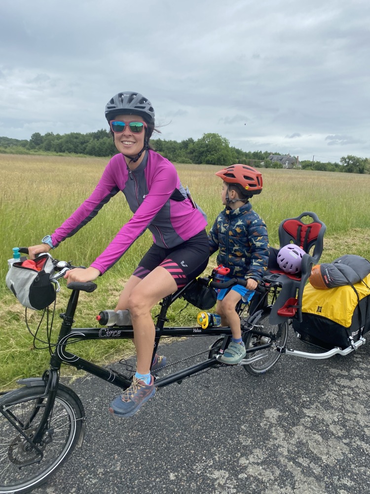 the yellow and black Burley Nomad cargo trailer hitched to a tandem with a mum, son and baby on board smiling infront of a green field