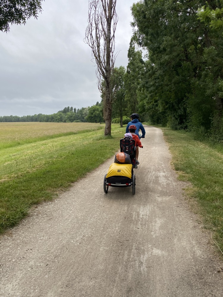 the yellow and black Burley Nomad cargo trailer hitched to a tandem with a mum, son and baby on board smiling infront of a green field