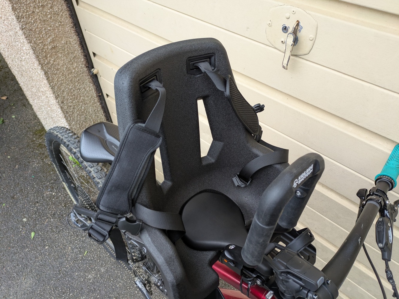 A Kids Ride Shotgun Pro Evo reat mounted to a red mountain bike leant against a garage door- a close up of the straps