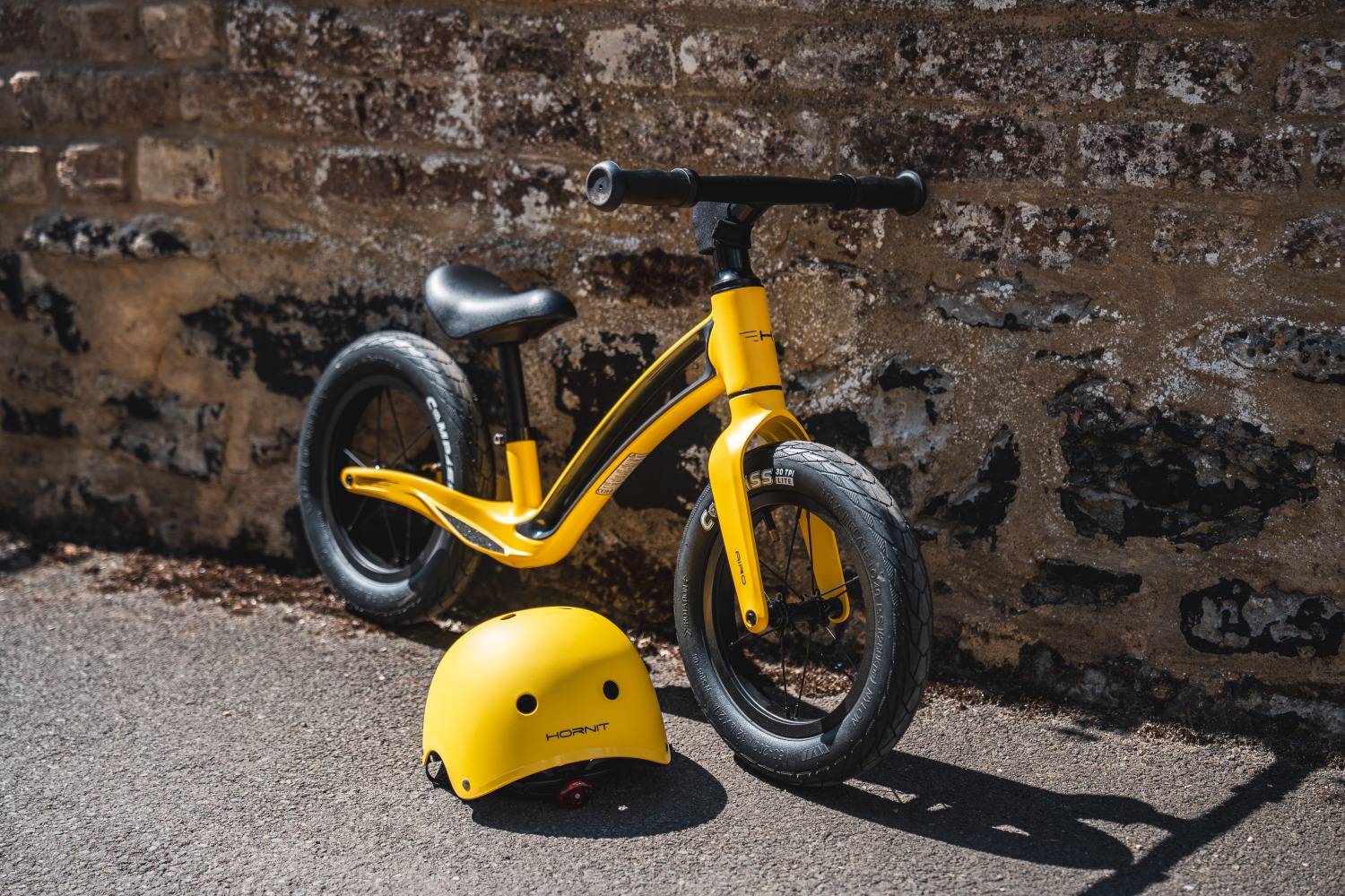 Yellow Hornit AIRO balance bike learning against a wall, with a yellow helmet next to it 