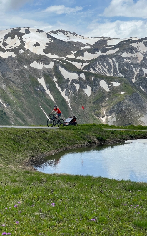 Hetty Kingston seen from the side and a distance, riding her bike and trailer up a smooth tarmac path with Swiss mountains in the background and a lake in the foreground