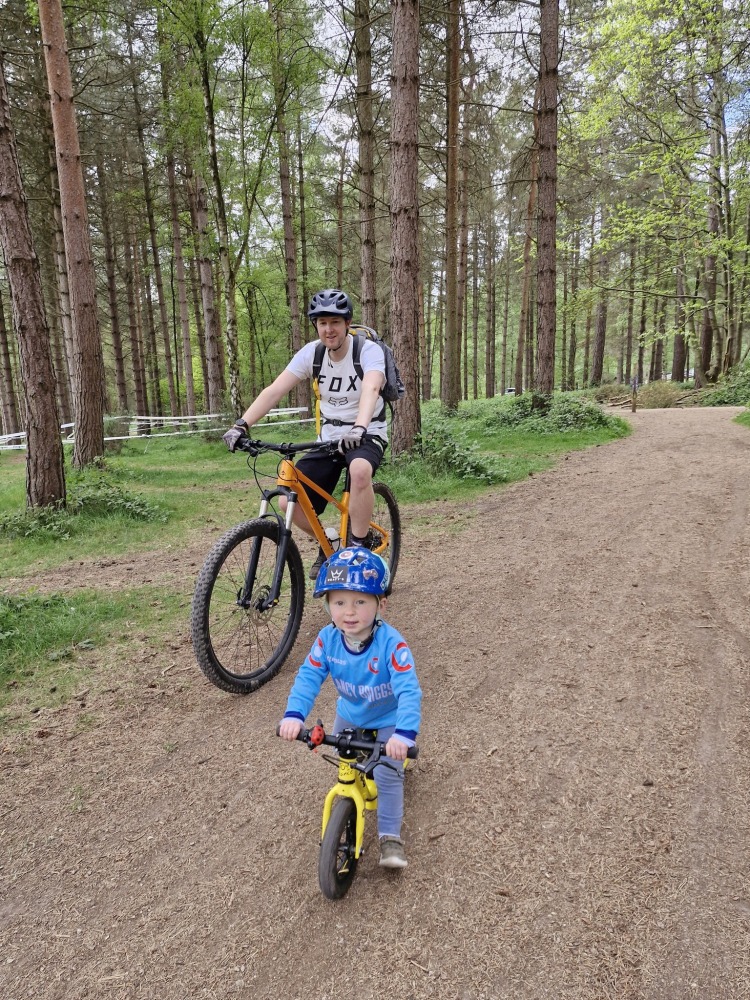A man riding a mountain bike alongside a small boy on a balance bike, with woodland in the background, seen from the front