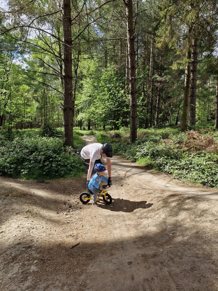 A dad bending over and helping his small son on his balance bike, in the middle of the woods