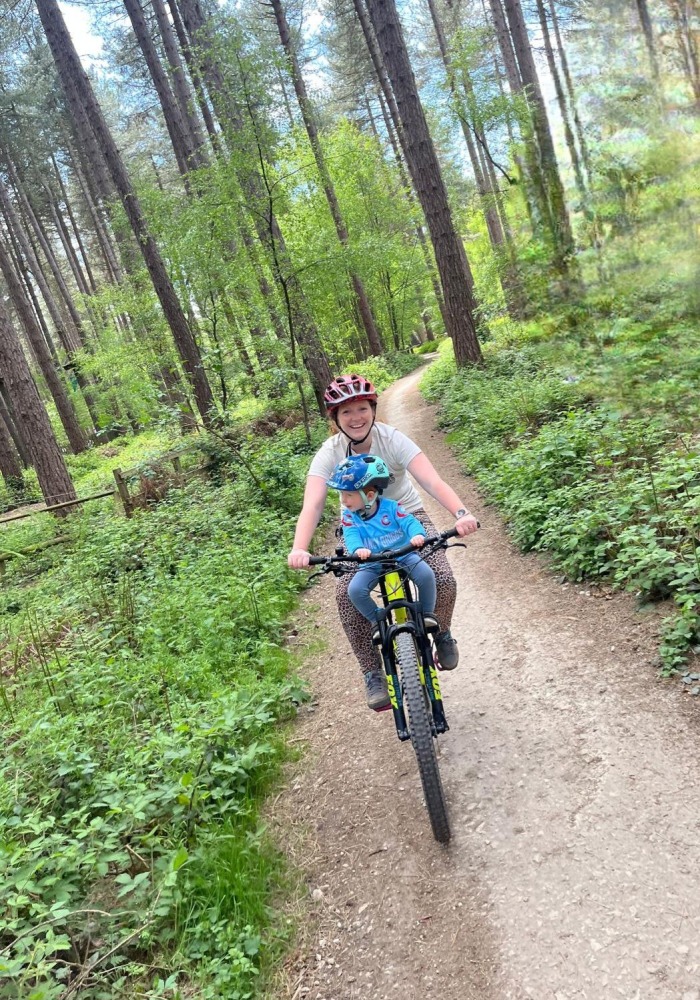 A woman seen from the front, riding a mountain bike through woodland, smiling at the camera, with her young son on a front bike seat