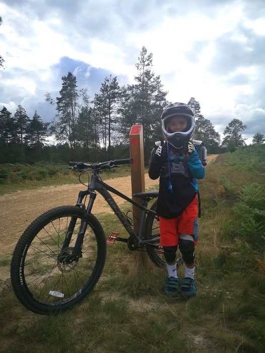 A little girl wearing a full face helmet standing next to her mountain bike and a red trail sign post, giving two thumbs up to the camera