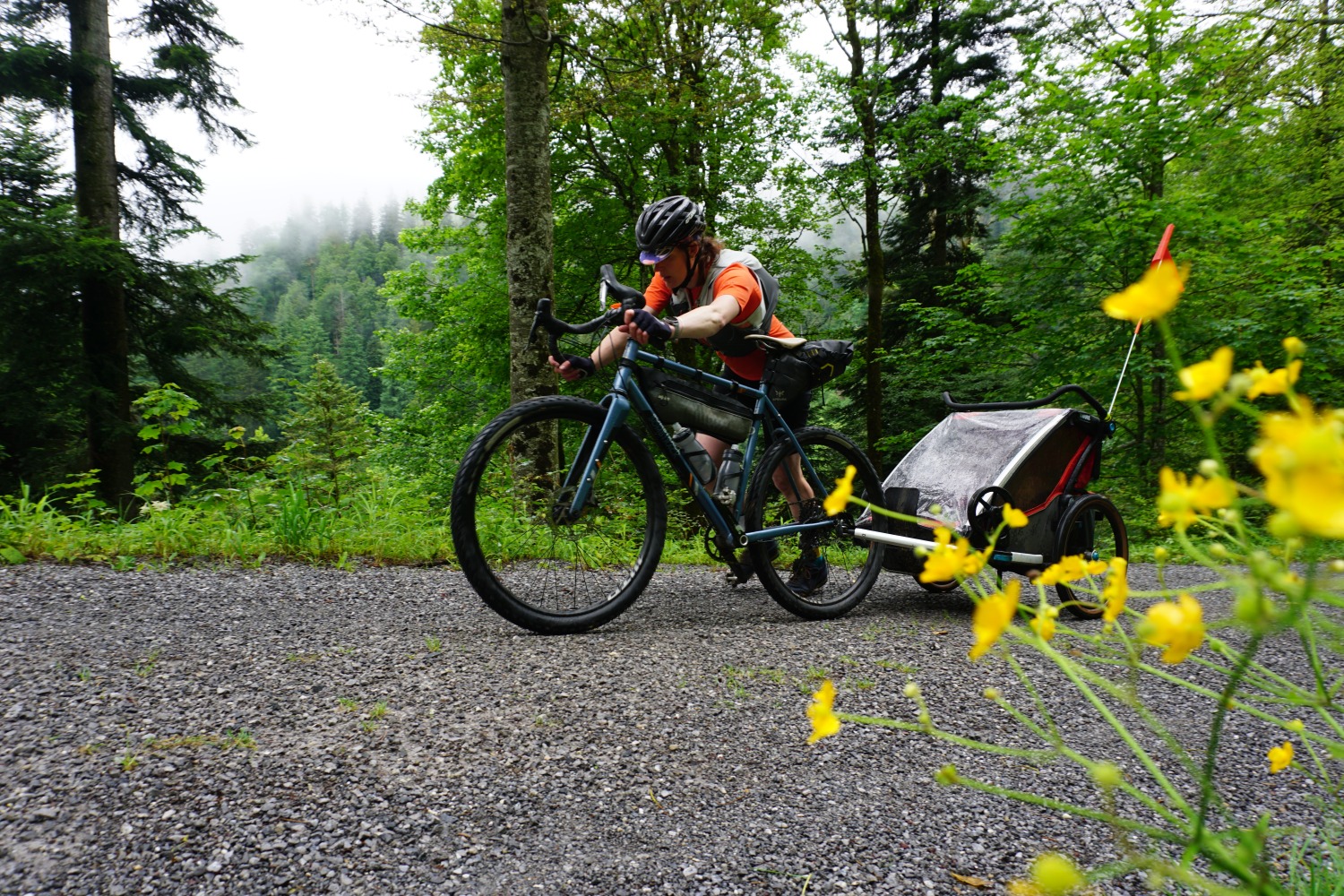 Hetty Kingston seen from the side, pushing her bike on foot, with the twins in a trailer behind her, trees in the background