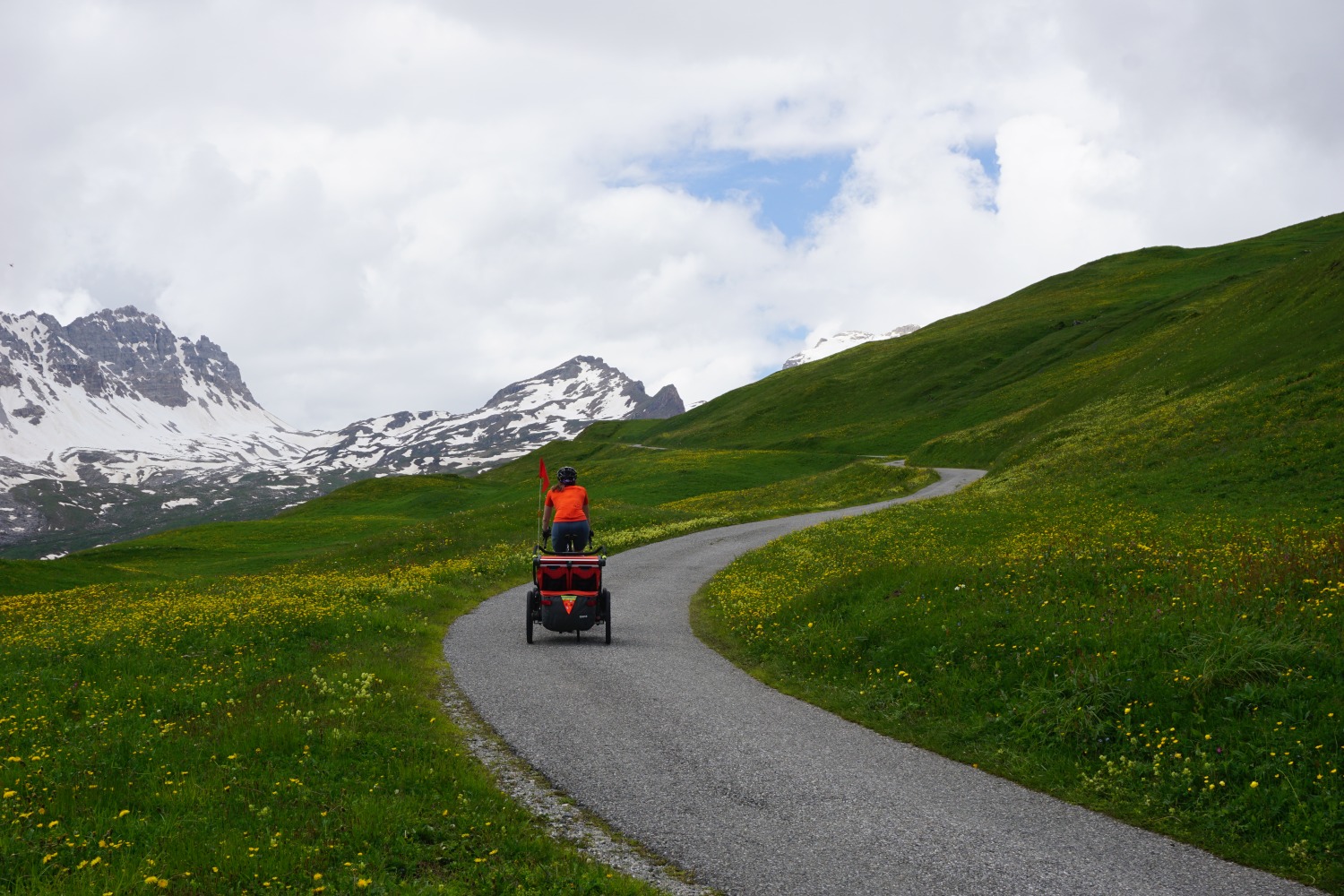 Hetty Kingston seen from behind cycling with the trailer along a smooth winding tarmac path with Swiss snow-topped mountains in the distance