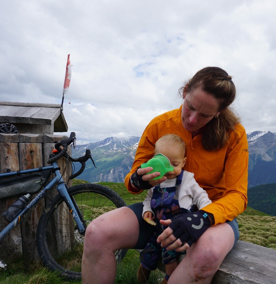 Hetty Kingston sat on a bench with her bike propped up next to her, and a toddler on her lap, who she's helping to drink from a sippy cup