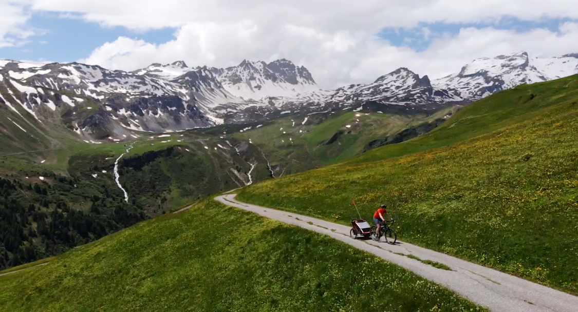 Hetty Kingston seen from a distance, cycling with the trailer along a tarmac path with Swiss snow-topped mountains in the distance
