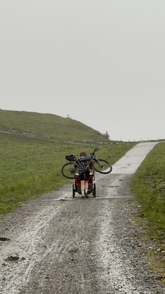 Hetty Kingston, seen from the front, cycling along a smooth tarmac road in rainy conditions, pulling the twins in a trailer behind her. The background is hazy with mist.