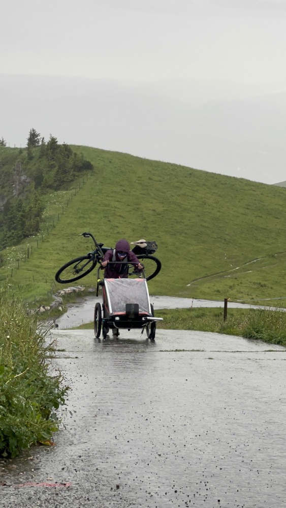 Hetty Kingston seen from a distance, carrying a bike on her back and pushing a kids trailer along a wet, smooth road
