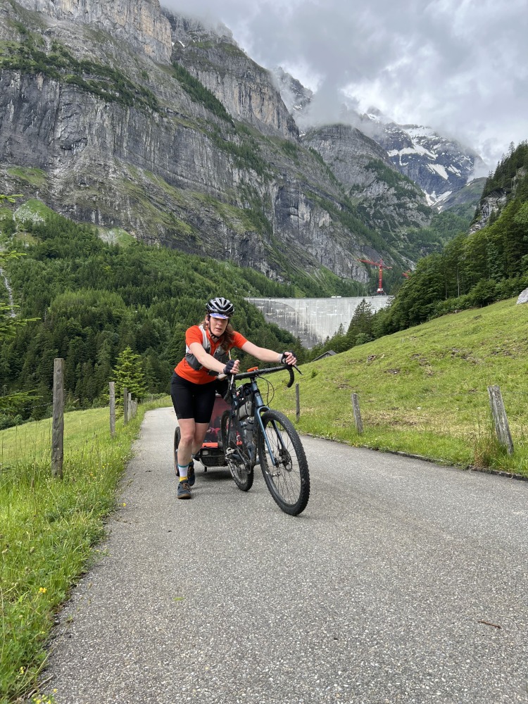 Hetty Kingston seen from the front, pushing her bike and trailer up a smooth tarmac path with Swiss mountains in the background