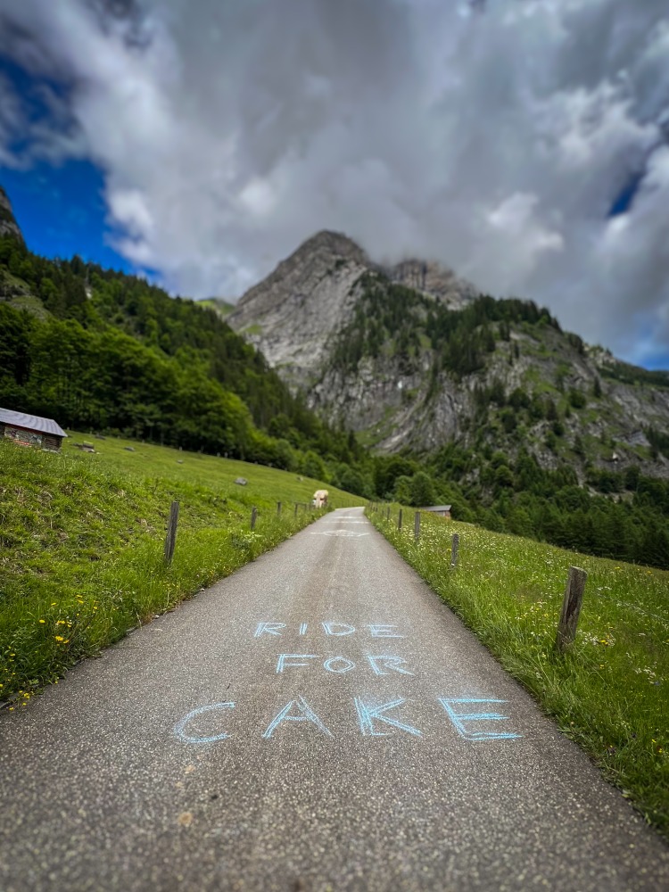 A tarmac road leading the mountains, with 'ride for cake' written on it in chalk
