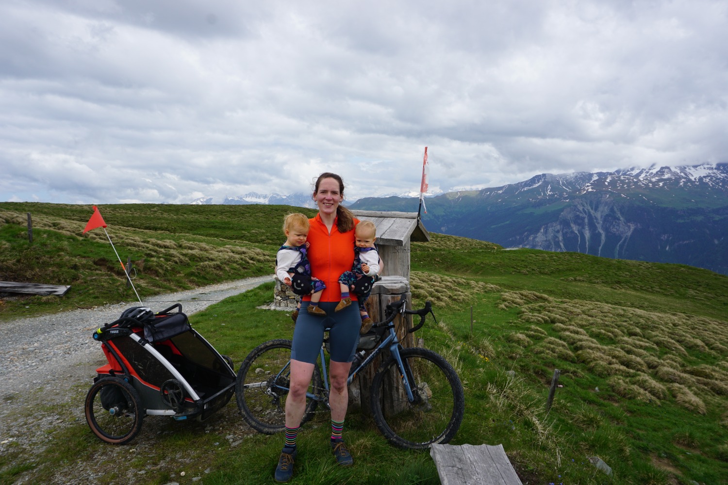 Hetty Kingston standing next to her loaded bike with a twin daughter in each arm and Swiss mountains behind her