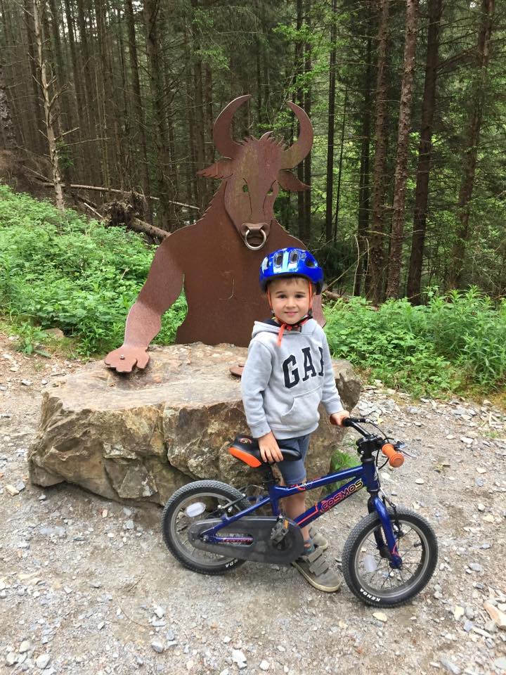 A little boy wearing a blue helmet posting with his bike in front of a Minotaur sign in the woods
