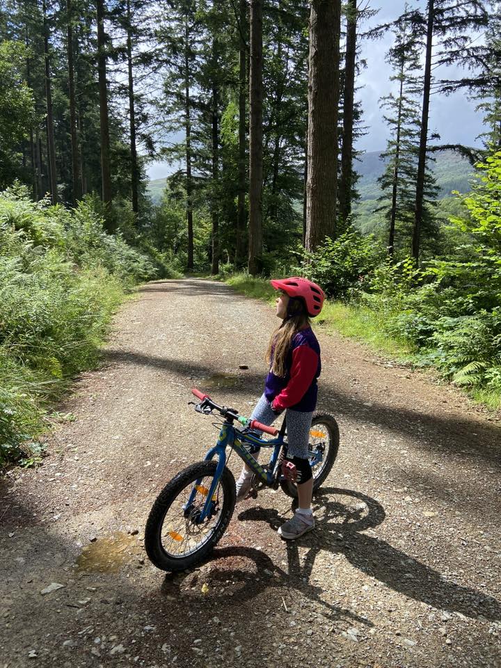 A little girl standing over her mountain bike on a gravel path surrounded by woodland, looking up at the trees