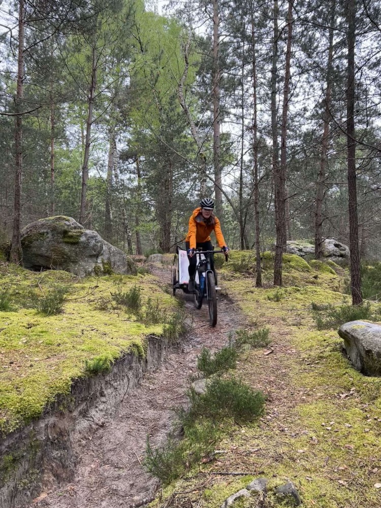Hetty Kingston cycling her bike while pulling a trailer, along a narrow rutted gravel path with trees behind her