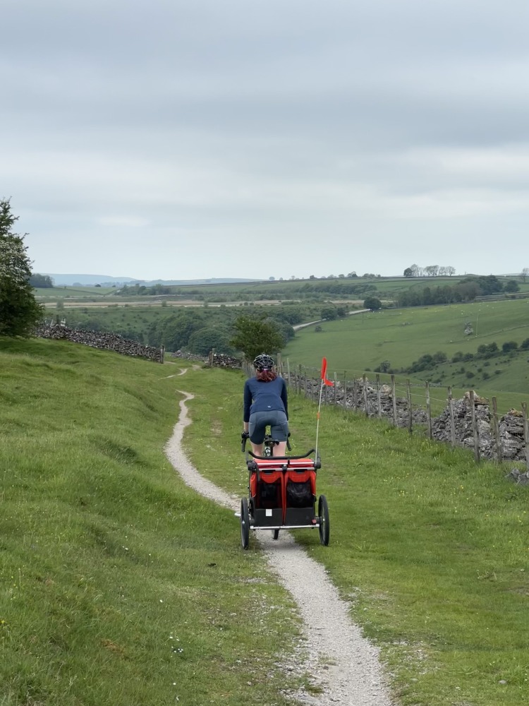 Hetty Kingston seen from behind, cycling and pulling a trailer along a very narrow gravel path surrounded by grass