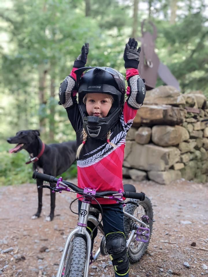 A little girl wearing a full face helmet, standing over a little bike and holding her hands in the air, with a dog and a Minotaur sign in the background