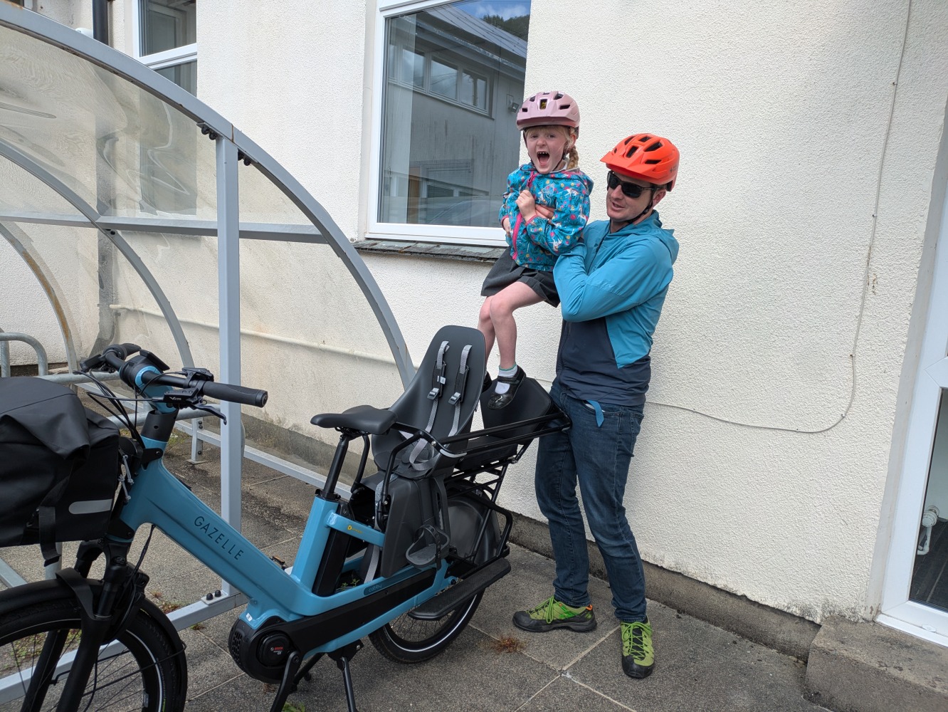 Gazelle Cabby review- a dad in a blue coat and orange helmet liftinghis daughter in a blue jacket and pink helmet and school dress into the bike